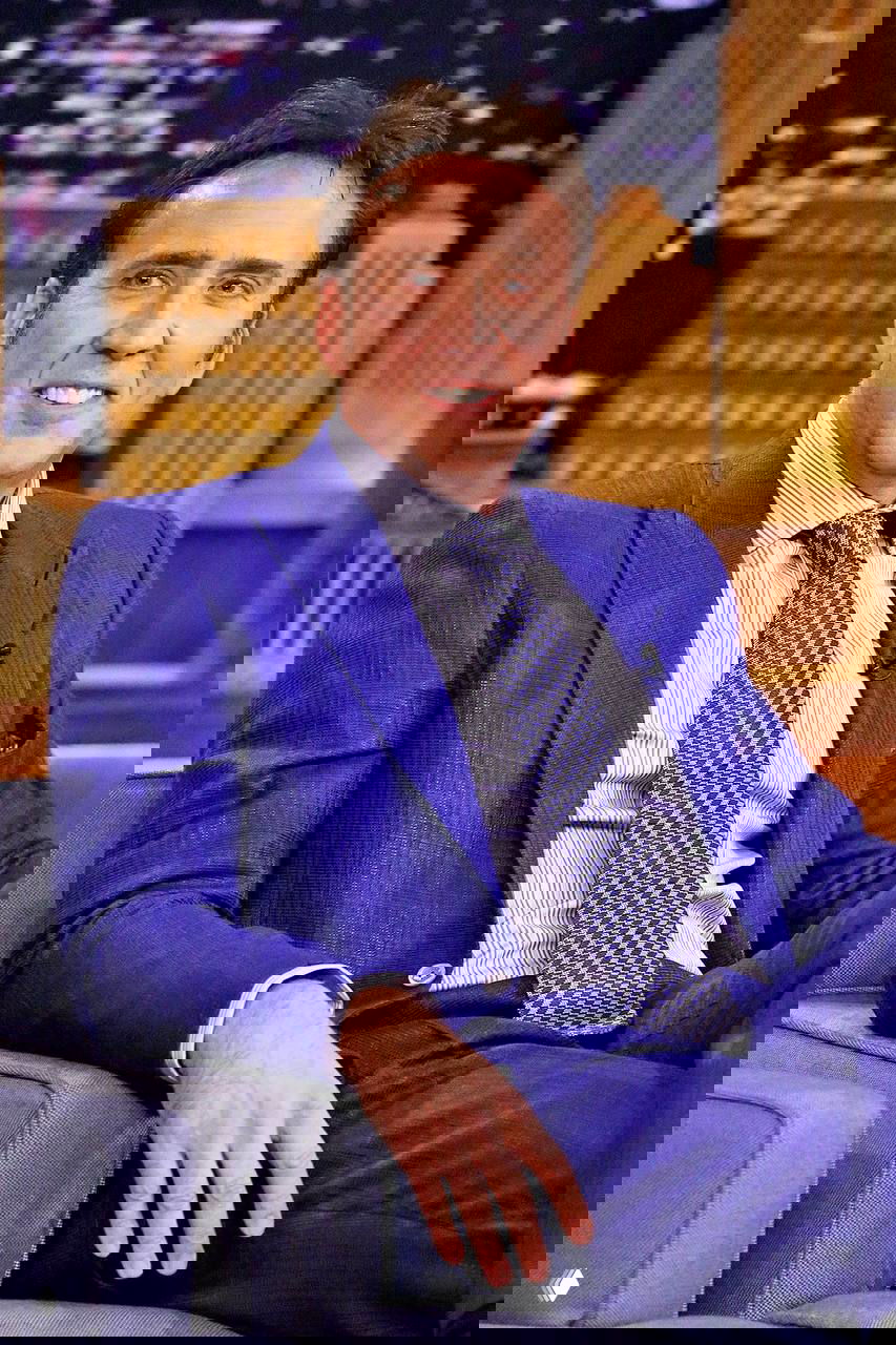 Герой в ярком костюме сидит в телевизионном кресле на ток-шоу, видны микрофон и студийный фон, деловой стиль одежды.