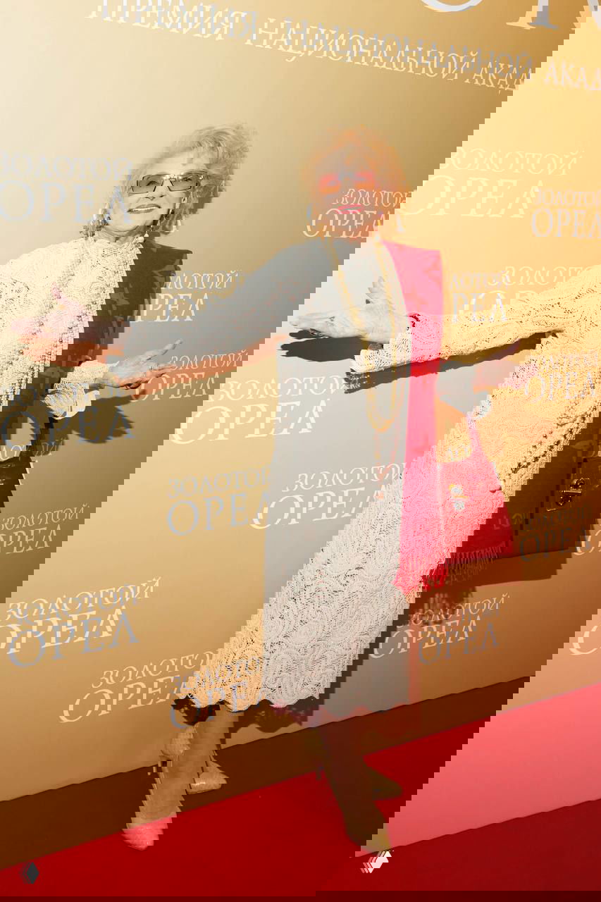 Светлана Дружинина в светлом кружевном платье и красном шарфе позирует на красной дорожке премии «Золотой орёл», улыбаясь фотографам.