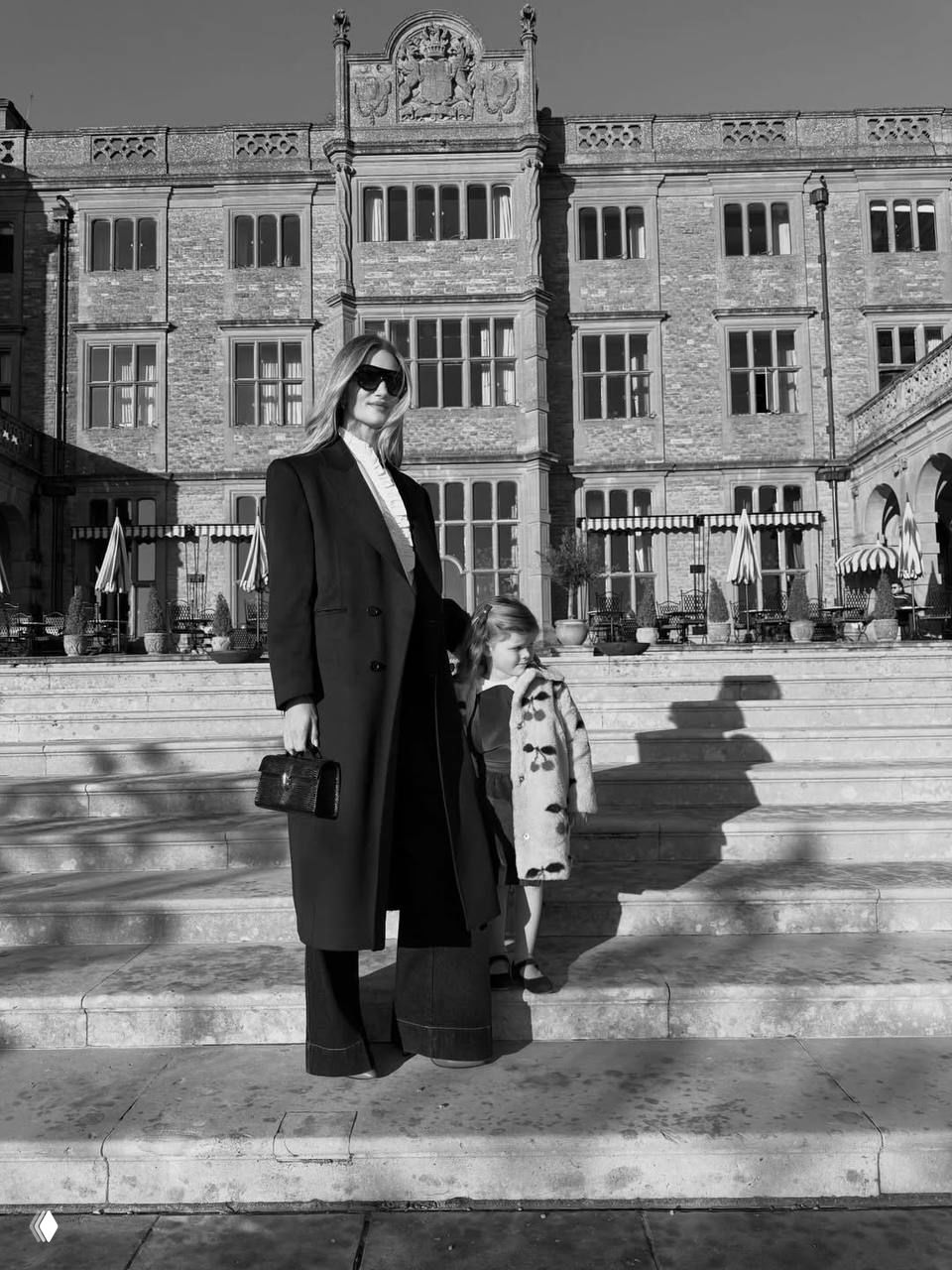 Чёрно‑белый снимок в полный рост: женщина в длинном пальто стоит на широких каменных ступенях рядом с ребёнком, заметна архитектура особняка на заднем плане