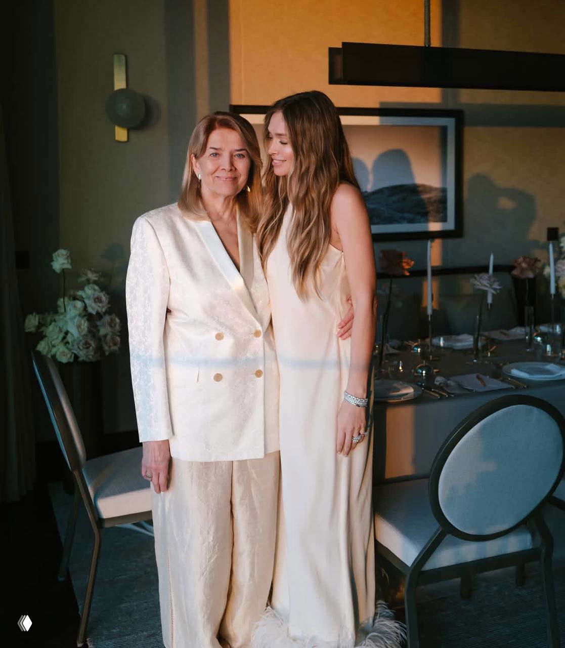 Две женщины в светлых нарядах стоят рядом у праздничного стола в ресторане: одна пожилая в жакете, другая — в длинном платье, обе в белой гамме.