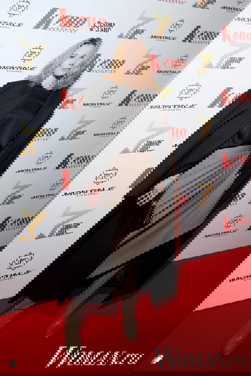 Женщина в светлом комплекте с пальто и юбкой, сапоги и клатч, позирует на фоне логотипов спонсоров и баннера «Кино Репортер».