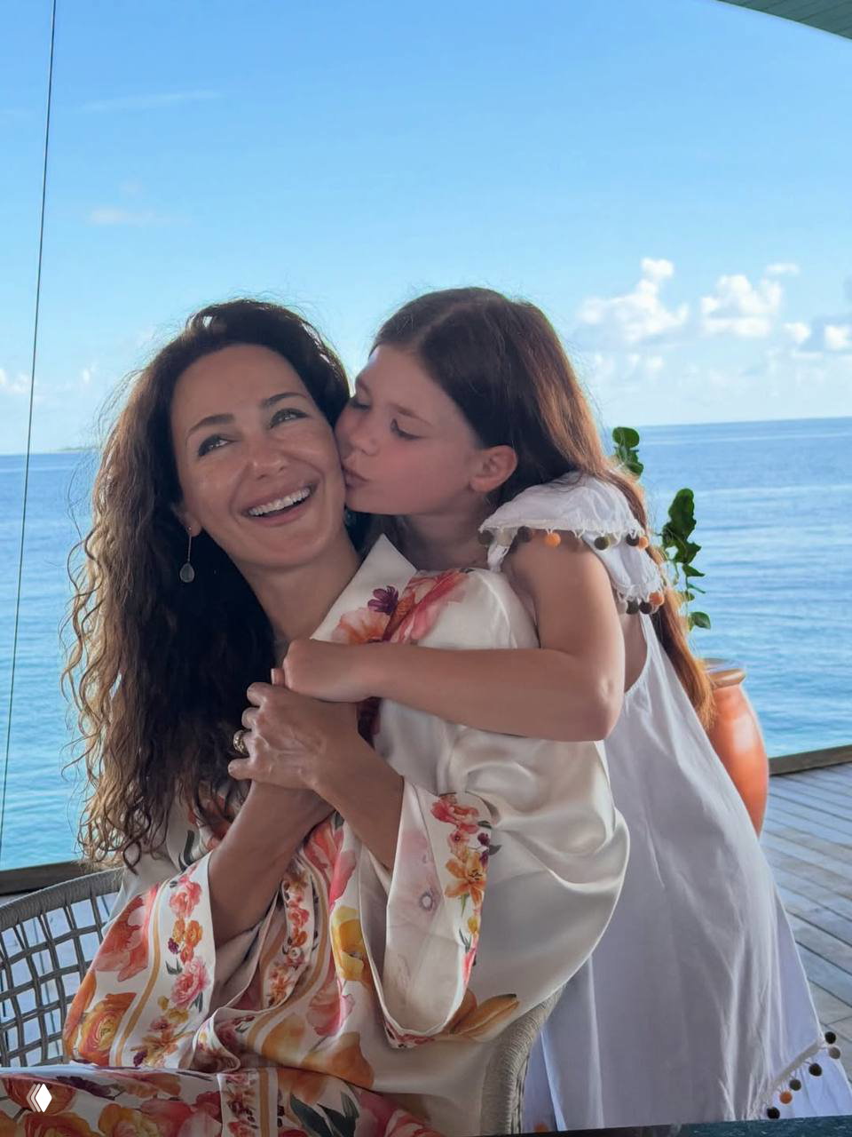 Сцена на открытой веранде у моря: женщина смеётся, девочка целует её в щёку; вдалеке спокойный океан и чистое голубое небо.