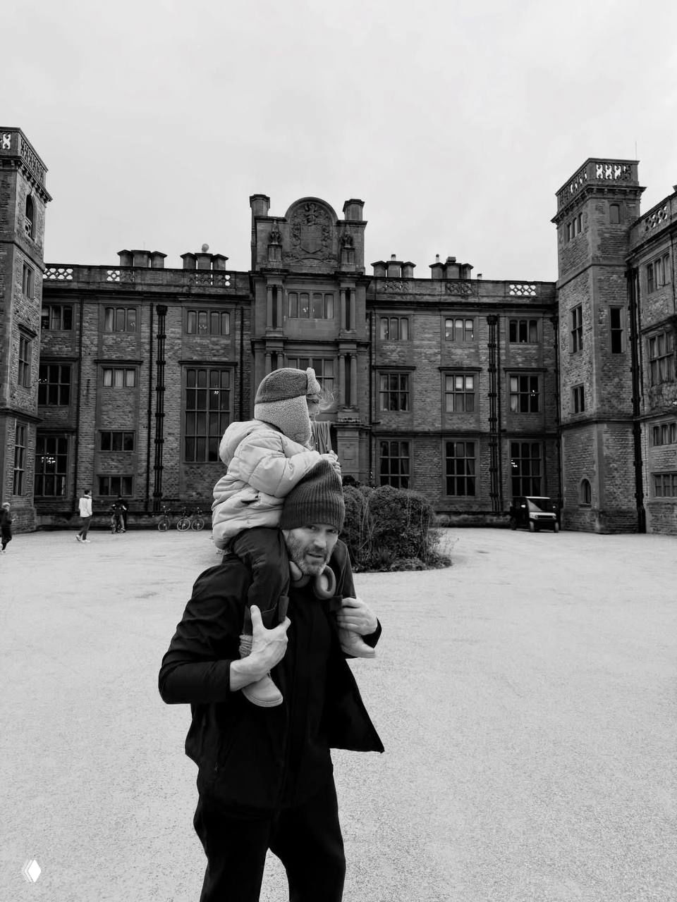 Чёрно‑белая фотография: мужчина несёт ребёнка на плечах по двору большого исторического особняка, динамичный семейный кадр с прогулки на открытом воздухе