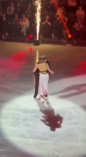Два фигуриста на льду во время шоу под прожекторами; одна спортсменка в белом платье с заметными пятнами на ткани и движением в центре круга света.