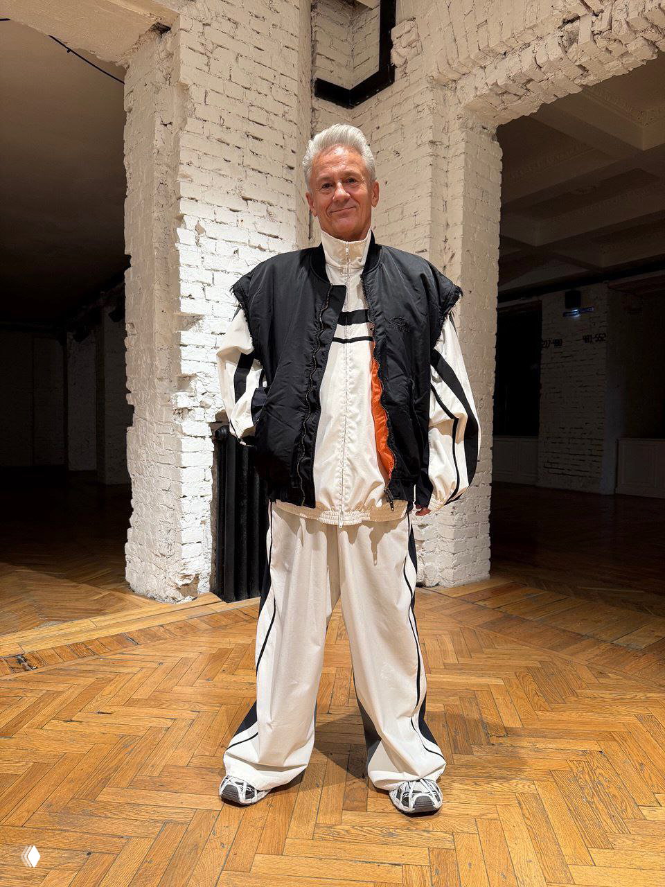 Актёр в спортивном светлом комплекте и тёмной стёганой жилетке: свободные брюки, кроссовки и просторная верхняя деталь на том же интерьерном фоне.