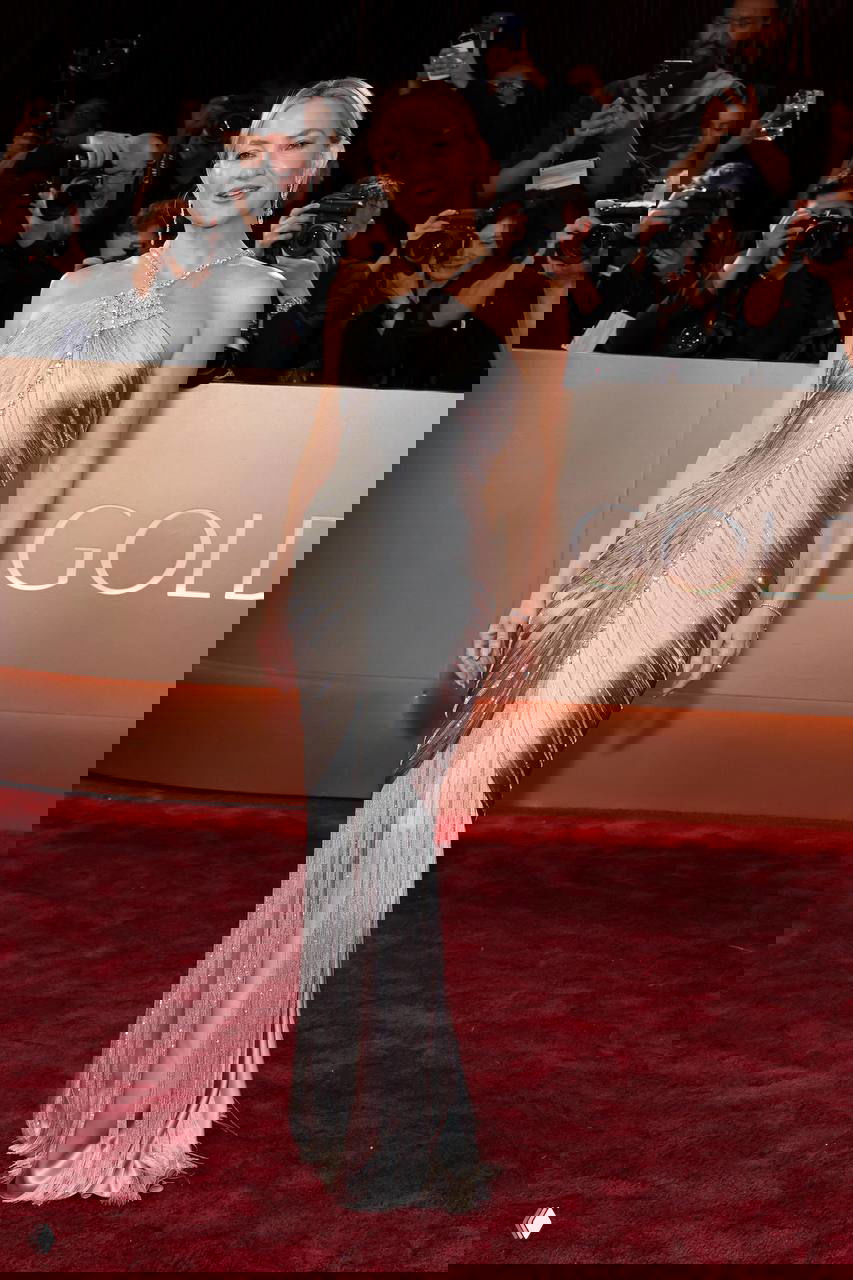 Женщина в серебристом облегающем платье позирует на красной дорожке «Золотого глобуса», в кадре фотозона и папарацци.