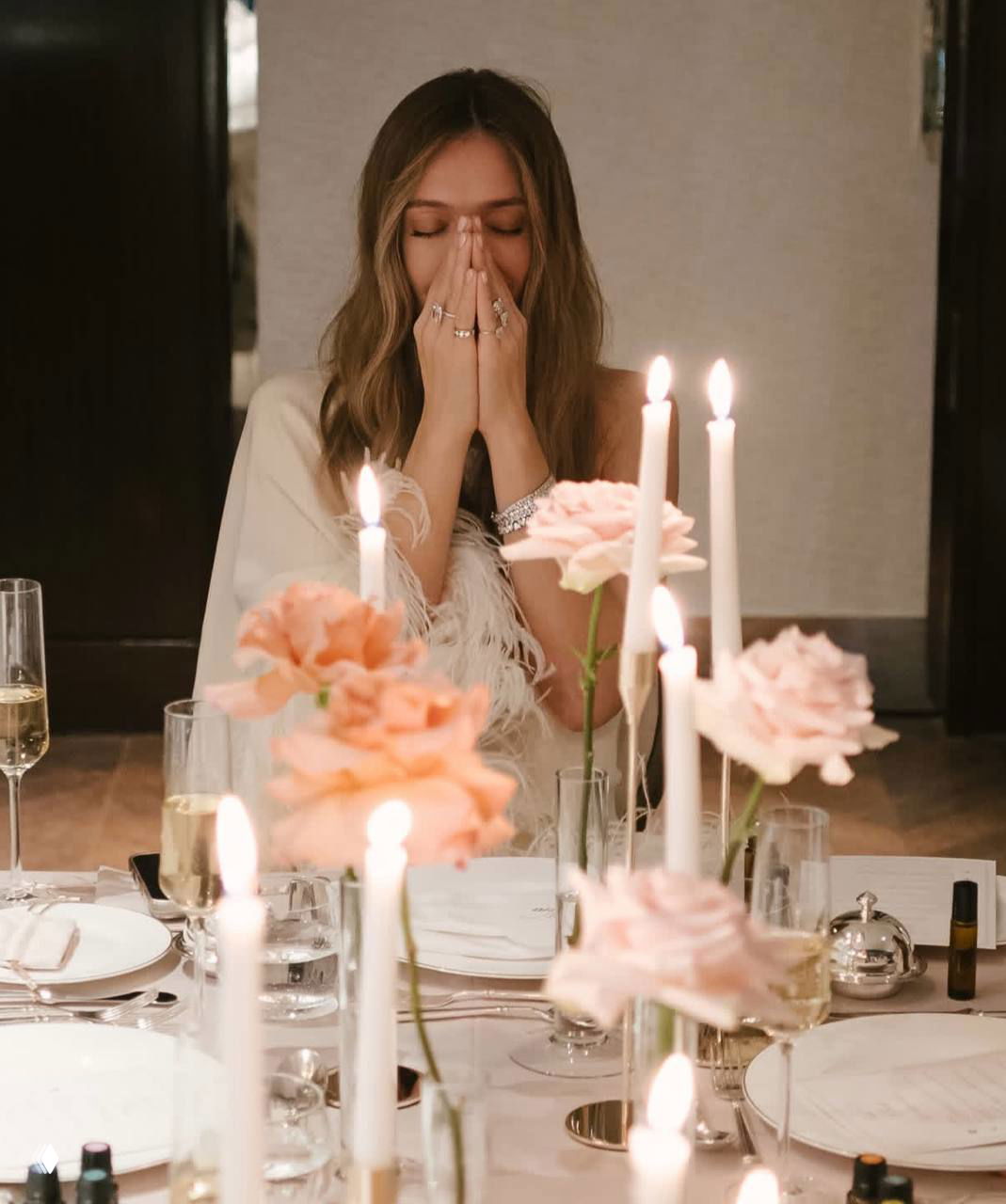 Женщина с закрытыми глазами сидит за сервированным столом с зажжёнными свечами и букетами роз в вазах на переднем плане праздничной композиции.
