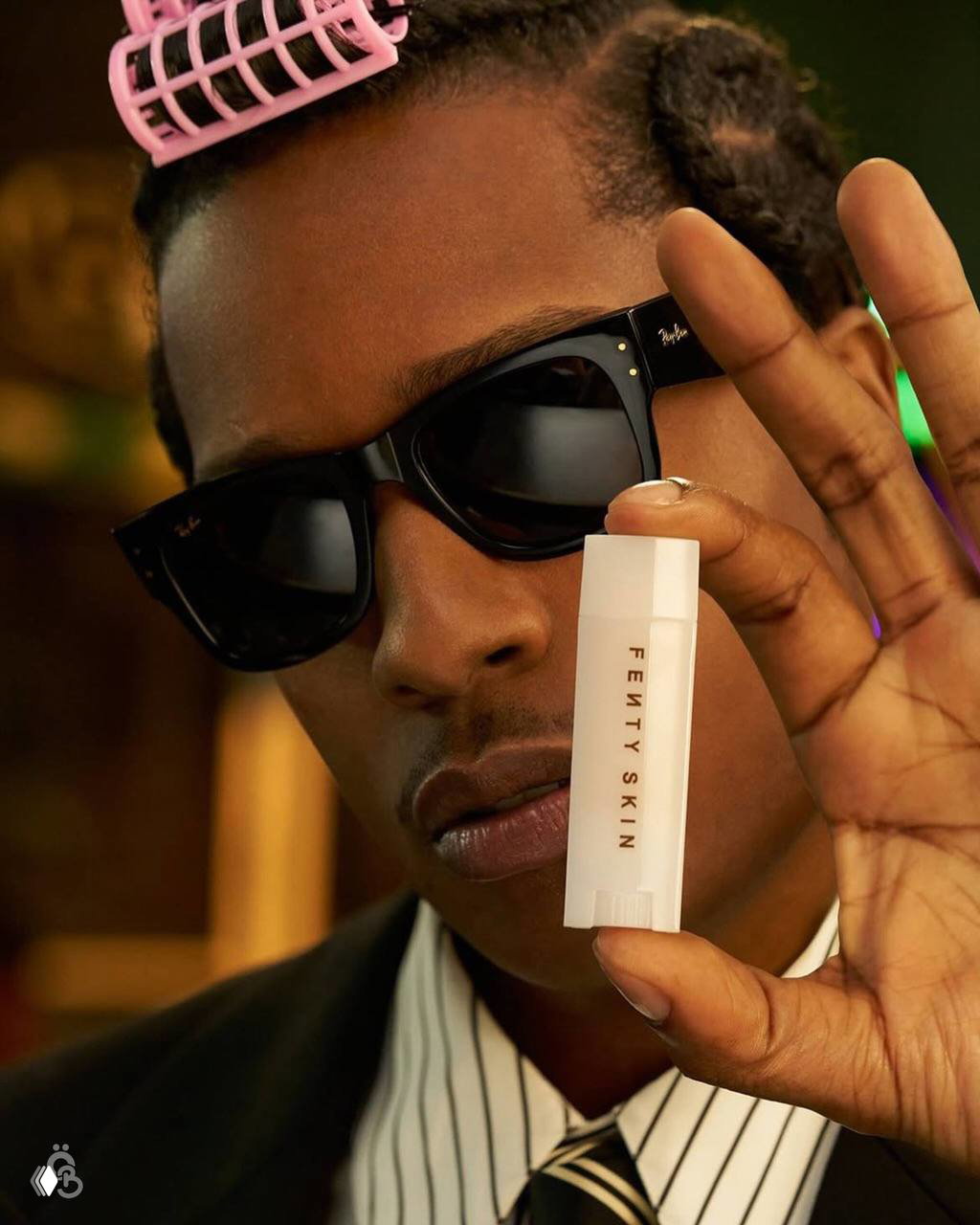 Крупный план лица A$AP Rocky в темных очках: он показывает белый бальзам для губ с надписью FENTY SKIN на фоне теплого студийного света.