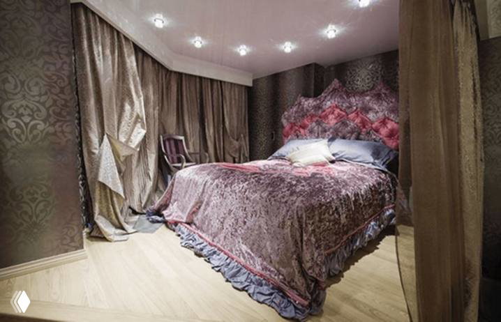 Сиренево‑фиолетовая спальня с пышной кроватью, драпированными тканями и декоративной спинкой; интерьер в богатой, почти королевской палитре и текстурах.