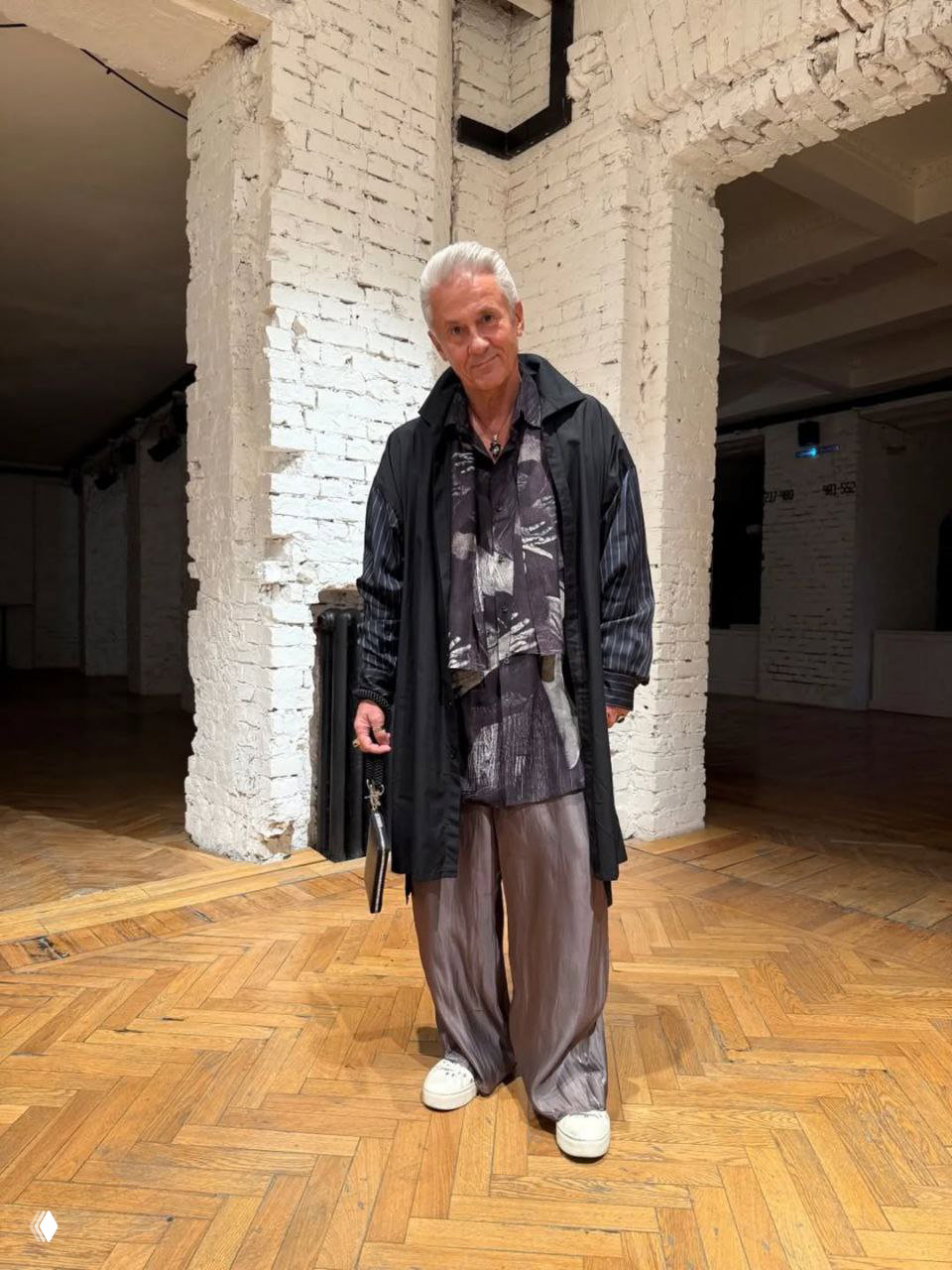 Мужской многослойный образ: тёмная рубашка с принтом, длинный плащ и свободные блестящие брюки на фоне белой кирпичной стены и паркетного пола.