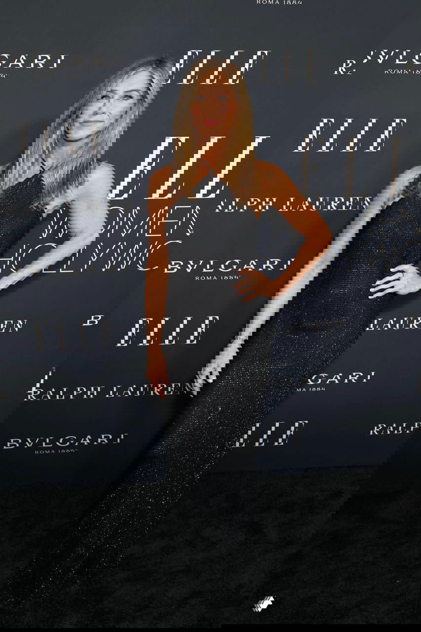 Дженнифер Энистон на церемонии ELLE Women in Hollywood в чёрном облегающем платье, позирует в полный рост на фоне логотипов мероприятия.