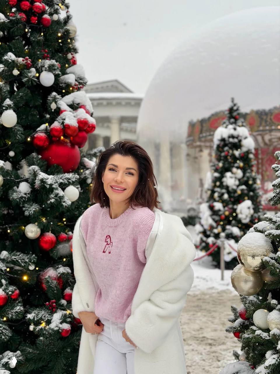 Женщина на зимней прогулке возле украшенной рождественской ёлки в пуховом белом пальто и розовом свитере, снежный городской фон и праздничная иллюминация