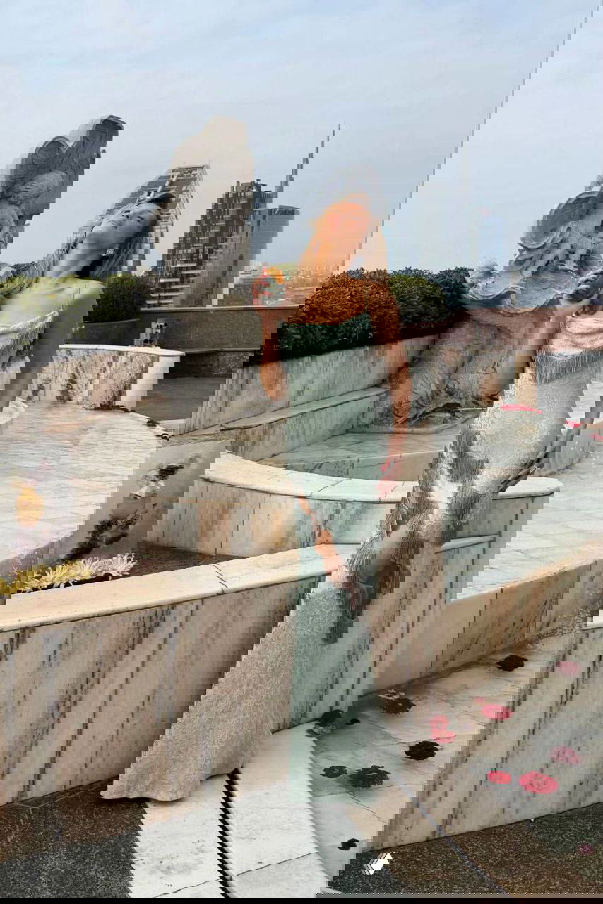 Белла Хадид у фонтанной скульптуры на террасе в платье Christian Dior, позирует с флаконом/аксессуаром Ôrəbella; архитектура и небо в кадре.