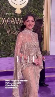 Улыбающаяся женщина в прозрачном блестящем платье на фиолетовой дорожке у фона с надписью Joy Awards и логотипом ELLE