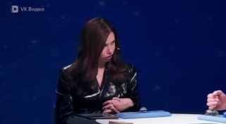 Кадр из студийного телешоу: женщина-ведущая в тёмном костюме сидит за столом, на столе папки и микрофон, тёмный фон.
