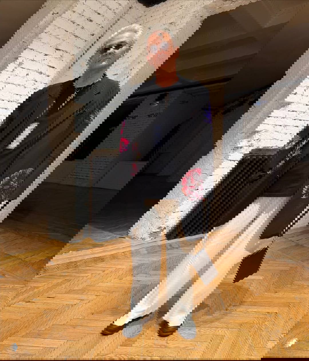 Олег Меньшиков в чёрном пиджаке с цветочными аппликациями, тёмной футболке и светлых свободных брюках — элегантный вечерний образ на фоне галерейного интерьера.