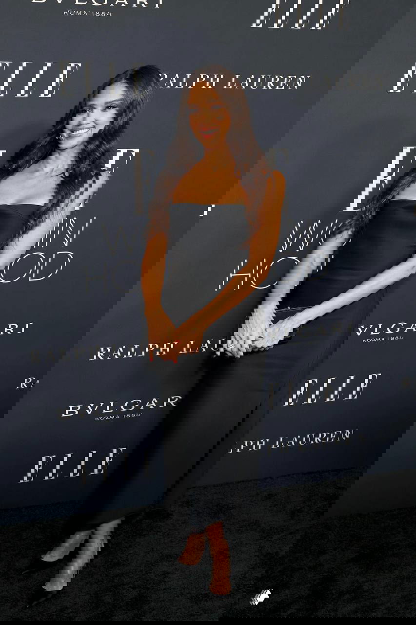 Джессика Альба на церемонии ELLE Women in Hollywood в чёрном открытом платье миди, улыбается фотографам на фоне фирменного баннера мероприятия.