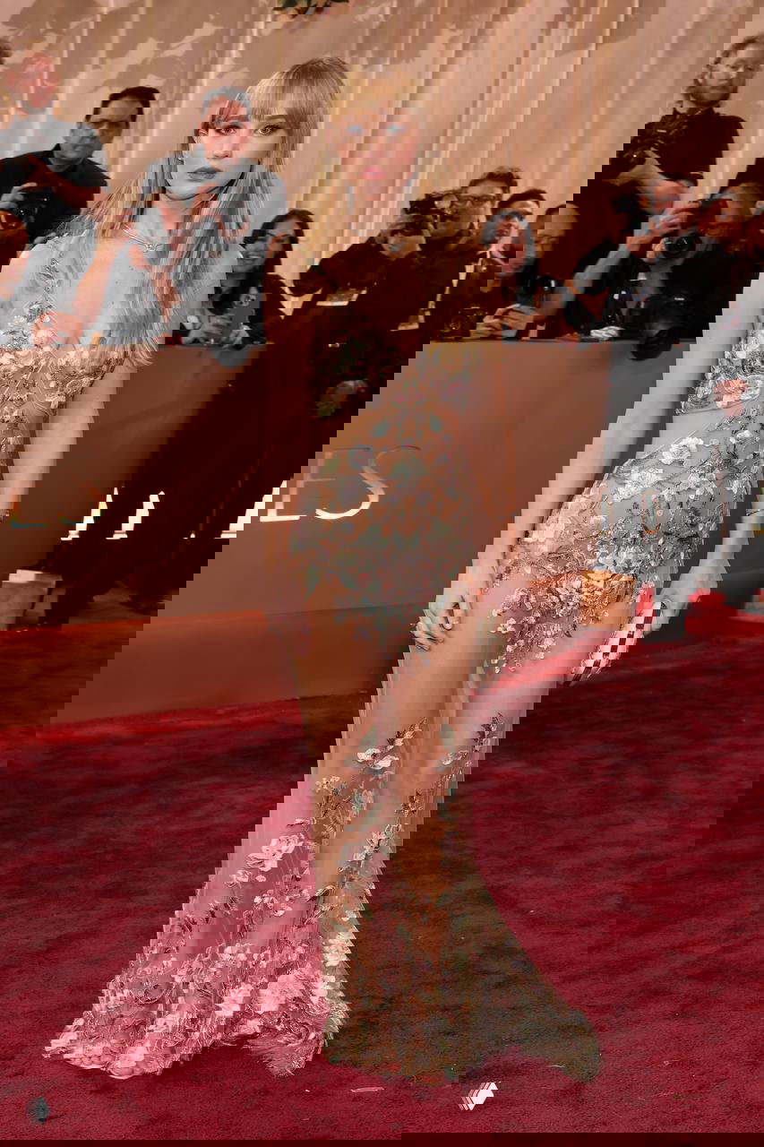 Женщина в прозрачном платье с цветочной вышивкой позирует на красной дорожке «Золотого глобуса», в кадре декор и фотографы.