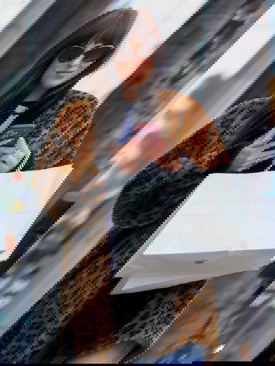 Крупный план: Дакота Джонсон держит телефон и пакеты, видна фактура леопардовой шубы и украшения на руках — повседневный стритстайл.