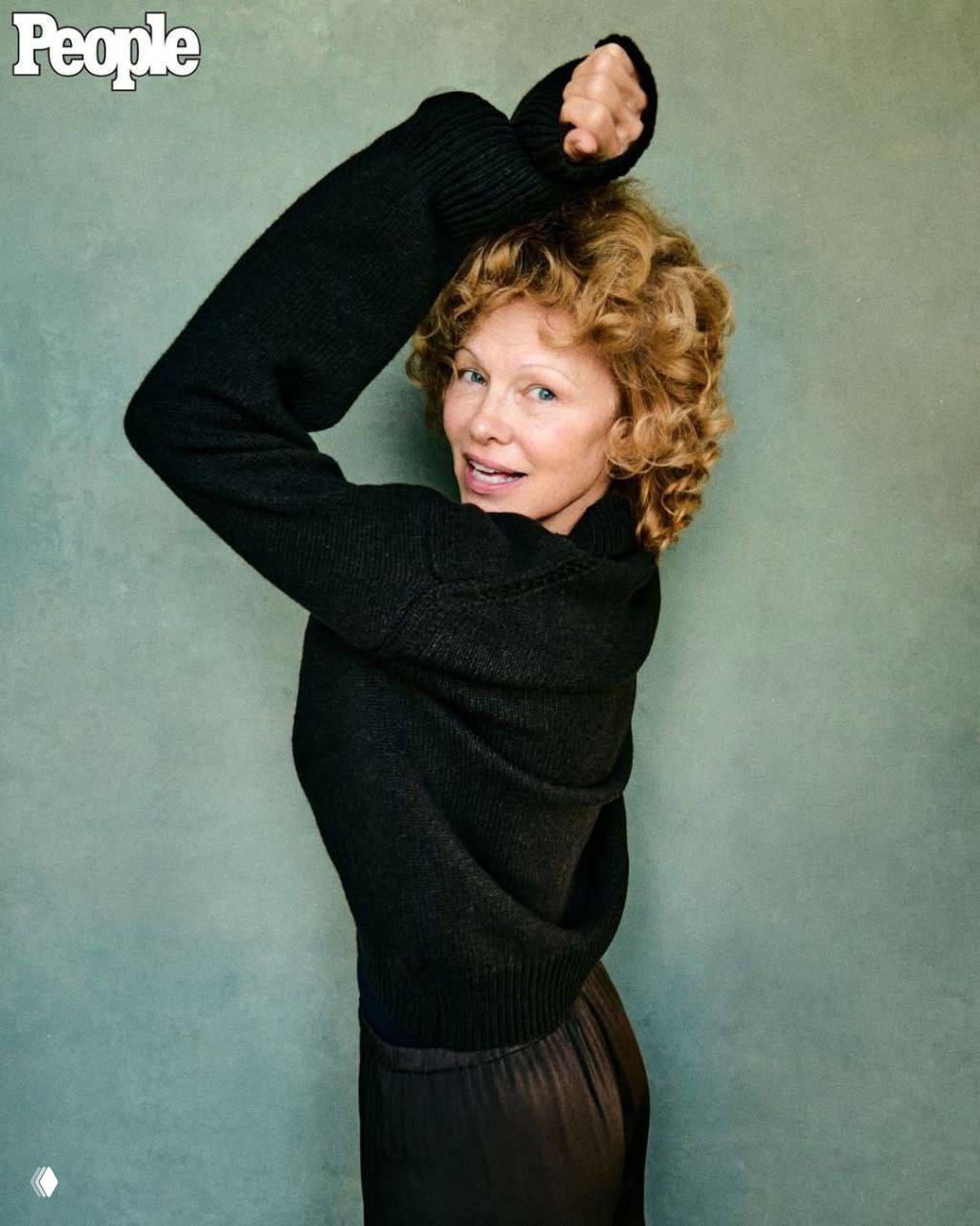 Памела Андерсон в полной фигуре на фоне однотонного фона в черном свитере, естественный макияж, поза в профиль и улыбка.