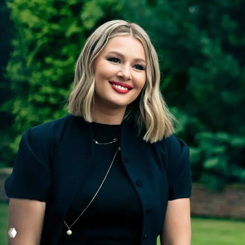 Портретный кадр на улице: женщина в деловой одежде улыбается на фоне зелёного парка — рекламный снимок для промо сериала.