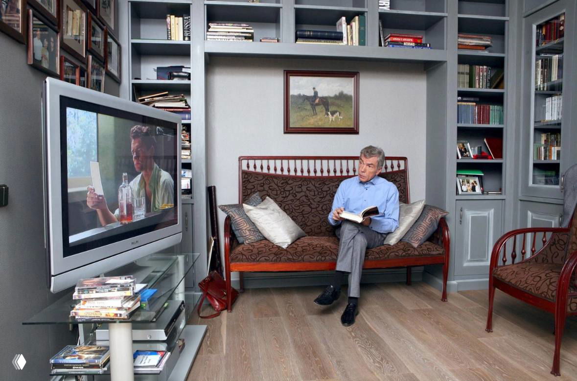 Телеведущий читает книгу в домашней библиотеке: диван, стеллажи с книгами и телевизор в интерьере квартиры на Саввинской набережной.