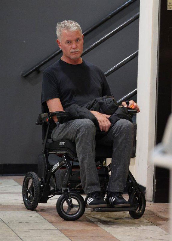 Актёр Эрик Дэйн в инвалидной коляске в помещении, в чёрной футболке и серых брюках, видно влияние болезни на подвижность.