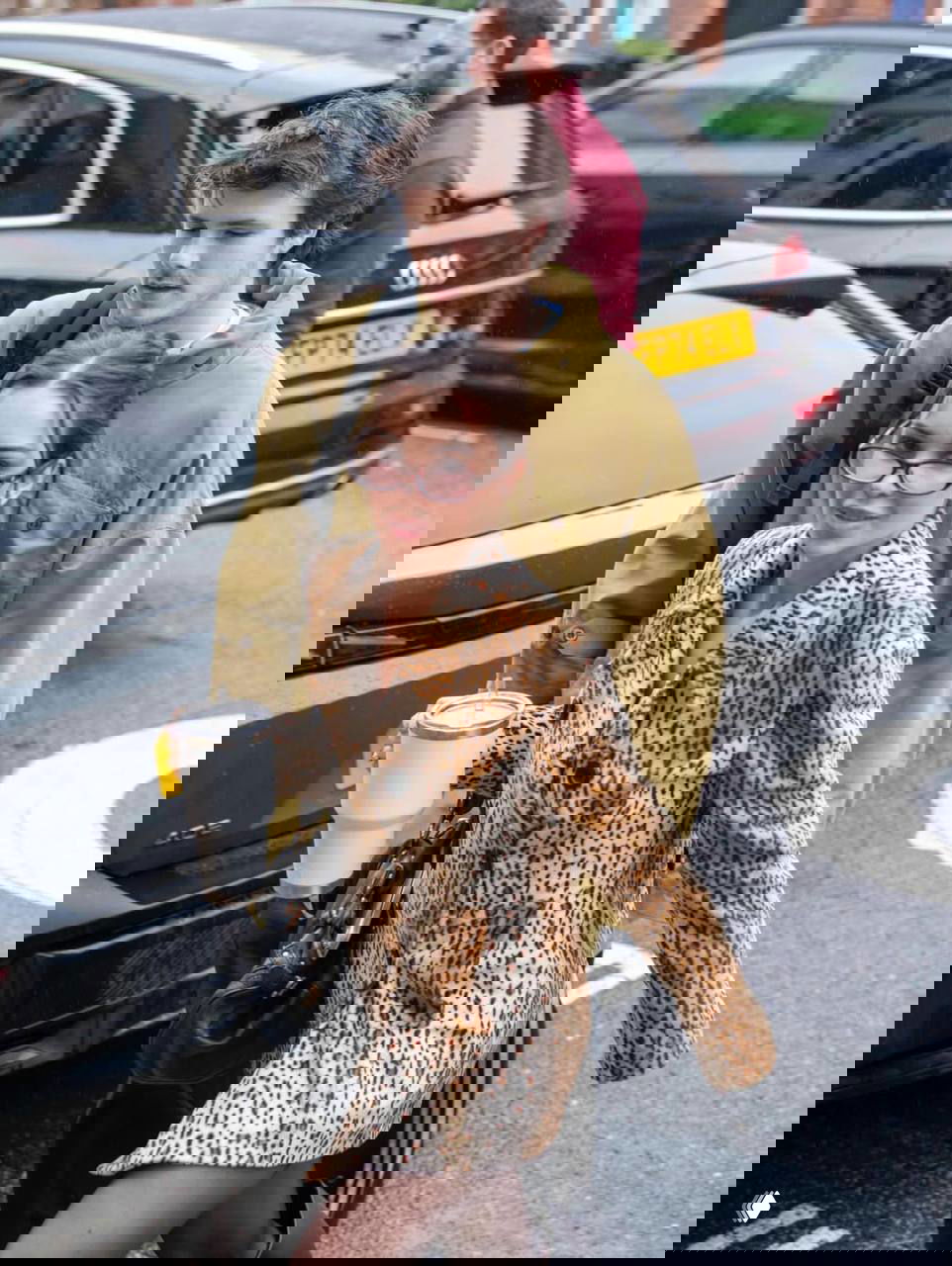 Джеки Апостел в коротком леопардовом пальто и очках держит термокружку, рядом идёт Круз Бекхэм в зелёной куртке; уличная сцена в дождливую погоду.