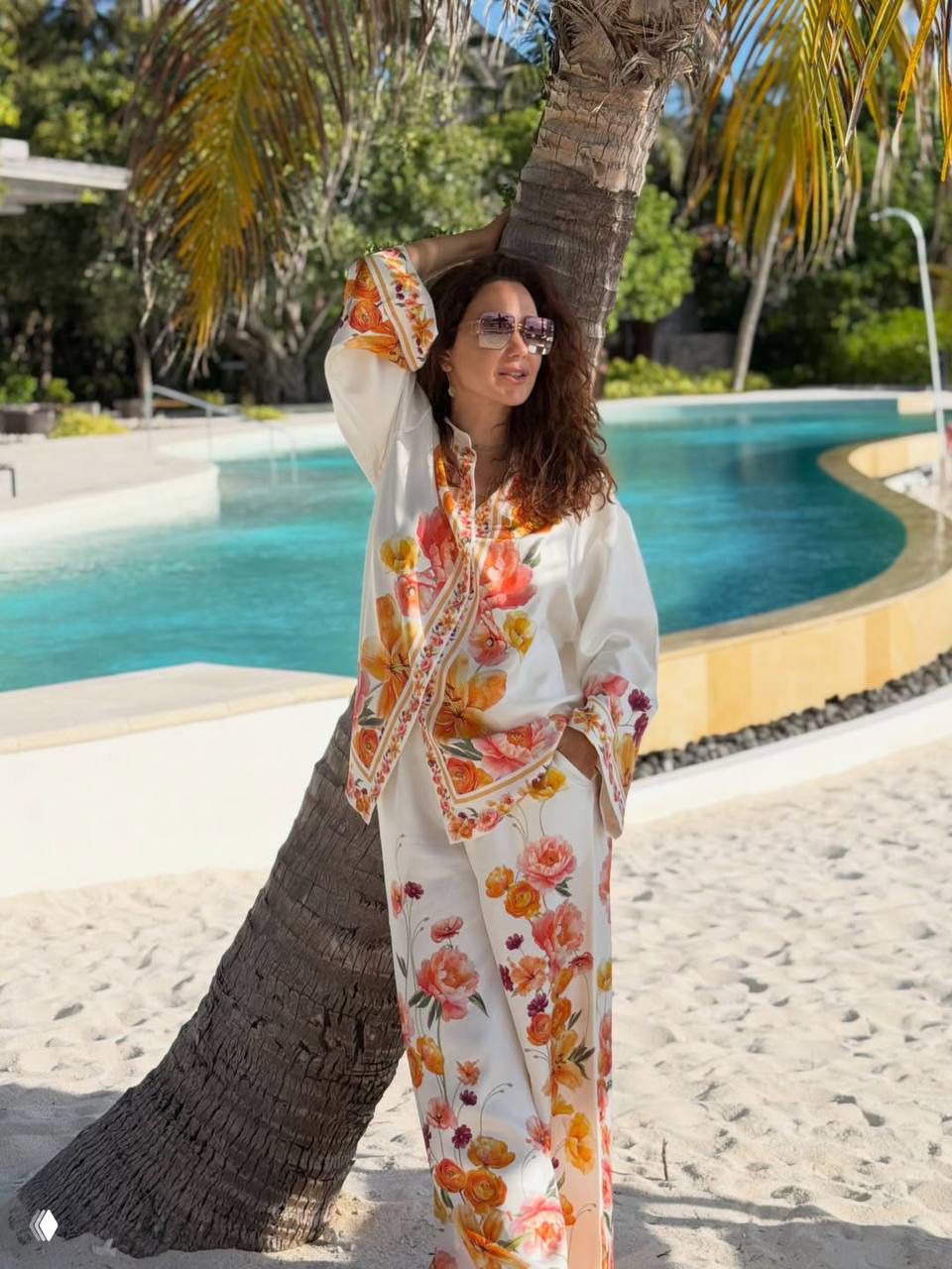 Женщина в цветочном костюме позирует у пальмы рядом с бассейном и песчаным пляжем; спокойная бирюзовая вода и зелень видны за ней.