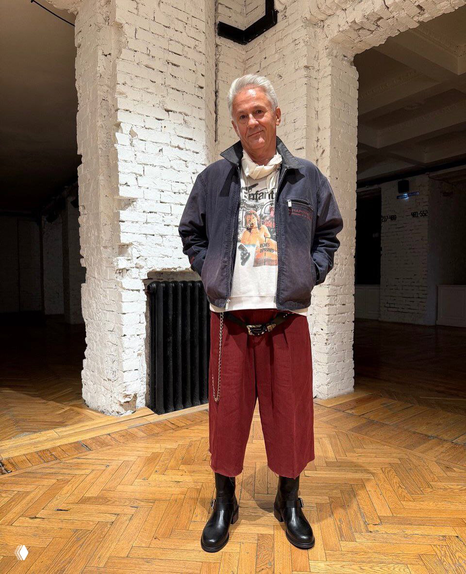 Кэжуал‑лук: укороченные бордовые брюки, белая футболка с принтом и тёмная куртка, дополненные ботинками и цепочкой на поясе.