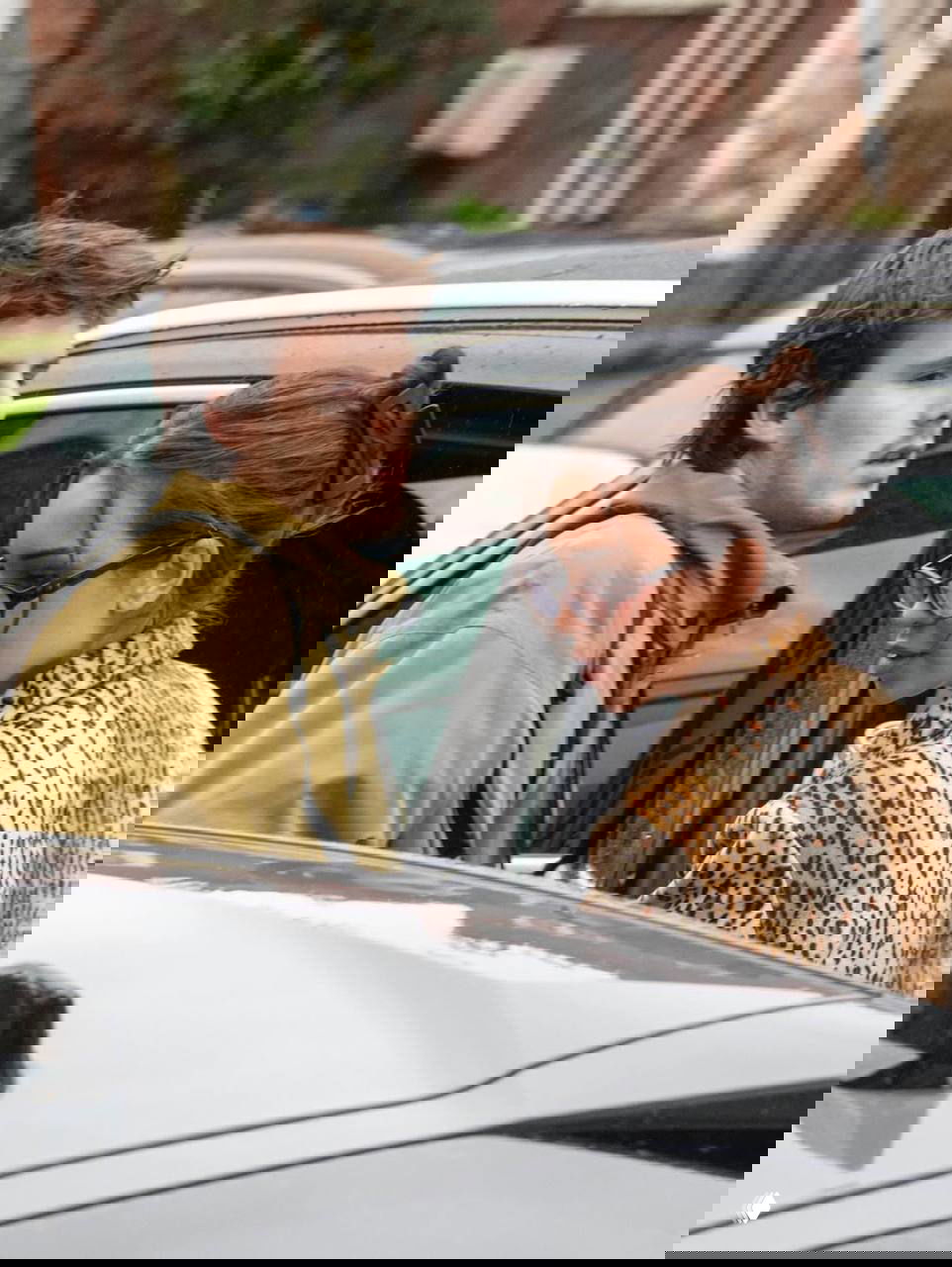 Крупный план: Джеки Апостел приближается к автомобилю, рядом Круз Бекхэм; кадр подчёркивает их совместный выход и взаимодействие с окружением.