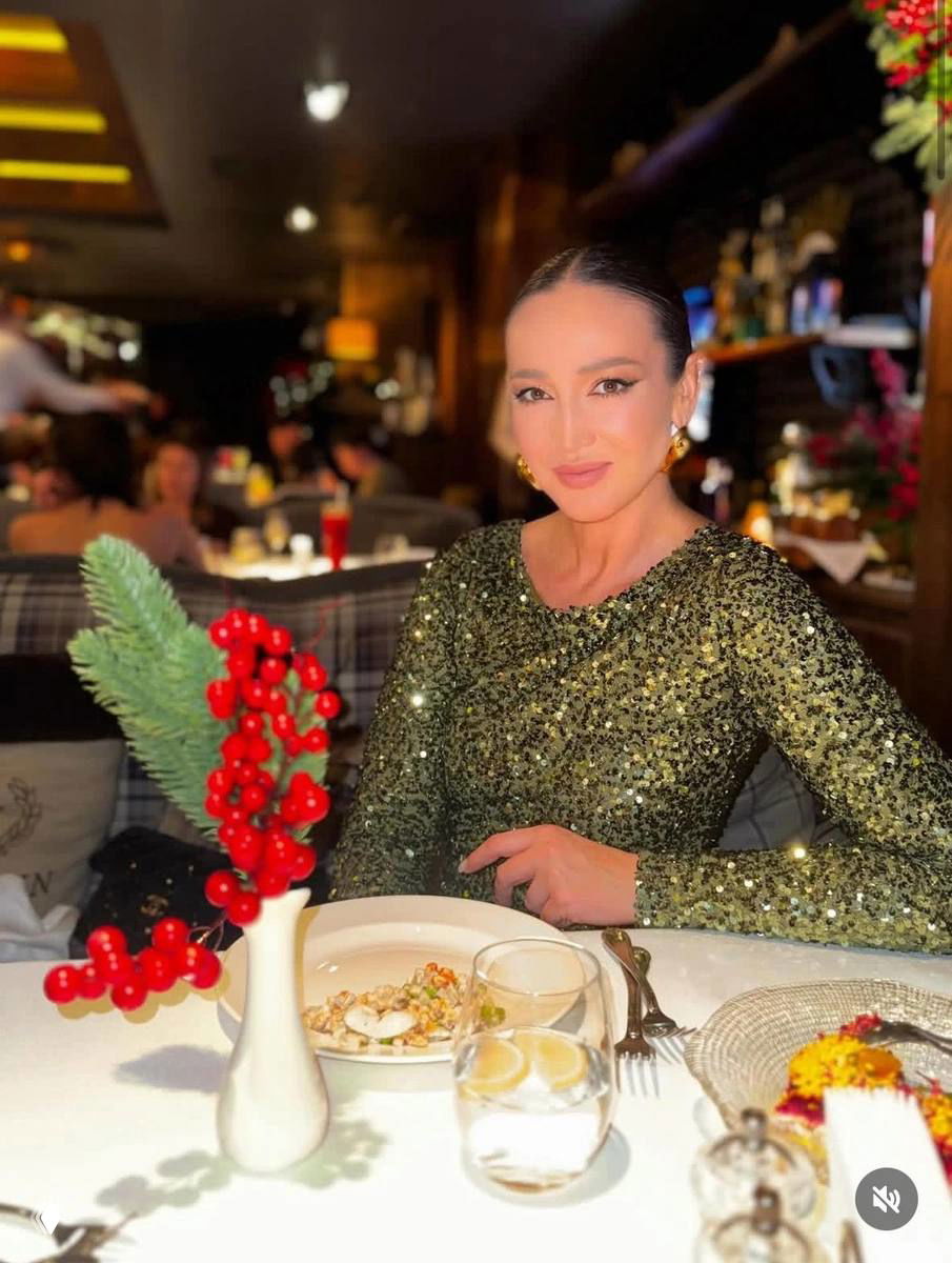 Женщина в блестящем платье сидит в ресторане за сервированным столом с рождественским декором, праздничная обстановка и тарелка с блюдом на переднем плане