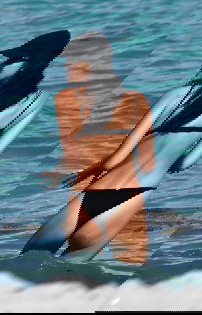 Женщина в чёрном бикини стоит в воде спиной к камере; видно обриси фигуры, морской пейзаж и ощущение пляжного отдыха.