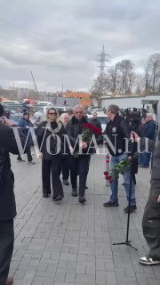 Группа людей идёт по тротуару с цветами и венками — посетители церемонии, которые пришли проститься с телеведущим.