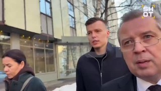 На фото трое людей идут по тротуару на фоне городского здания; зимняя одежда, на земле видны остатки снега и наледь, уличная сцена в городе.