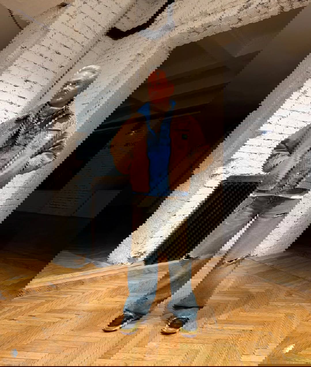 Слоистый деним‑лук: джинсы с потертостями, джинсовая рубашка и коричневая куртка поверх — мужской расслабленный стиль на паркетном полу.