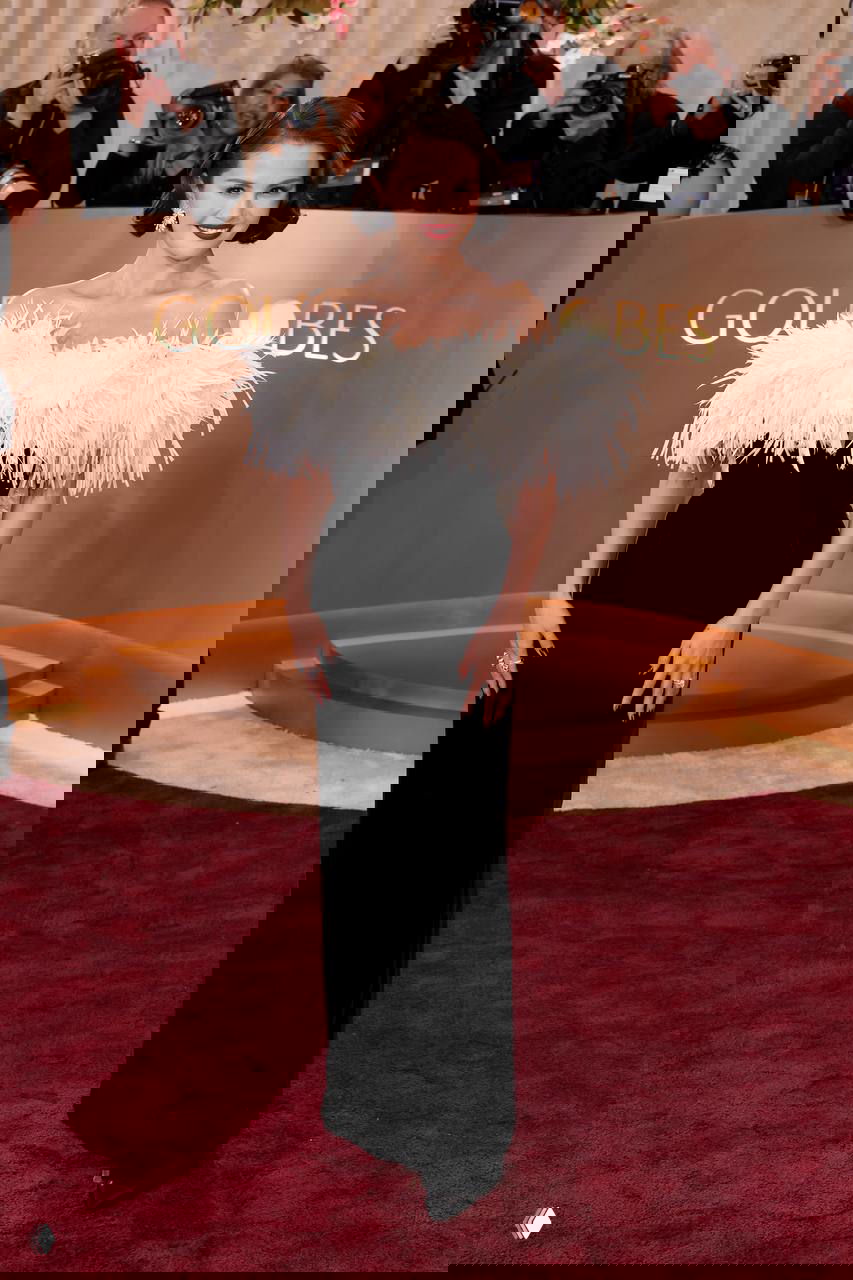 Женщина в чёрном платье с белым перьевым декором на линии плеч, красная дорожка и фотографы на заднем плане церемонии.