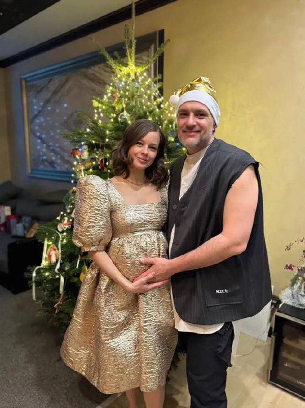 Будущая мама и мужчина позируют у украшенной ёлки дома: они держат руки на животе в тёплой семейной атмосфере, интерьер с рождественскими украшениями