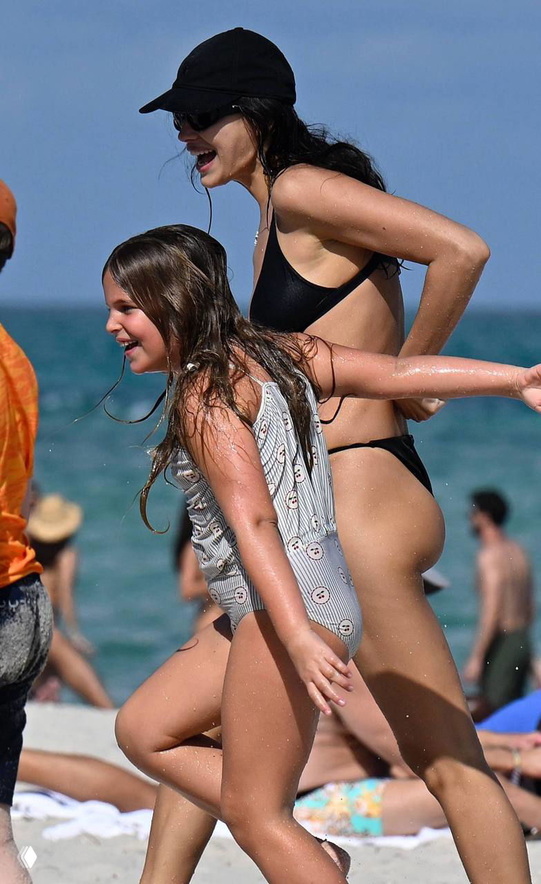 Женщина в чёрном бикини и девочка в цельном светлом купальнике бегут и смеются по песку, вместе играют у моря во время отпуска.