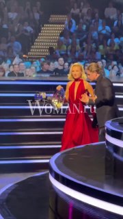 Полина Гагарина в красном платье выходит на сцену МУЗ‑ТВ 2026 с возлюбленным‑директором; на фоне зрители и логотип Woman.ru
