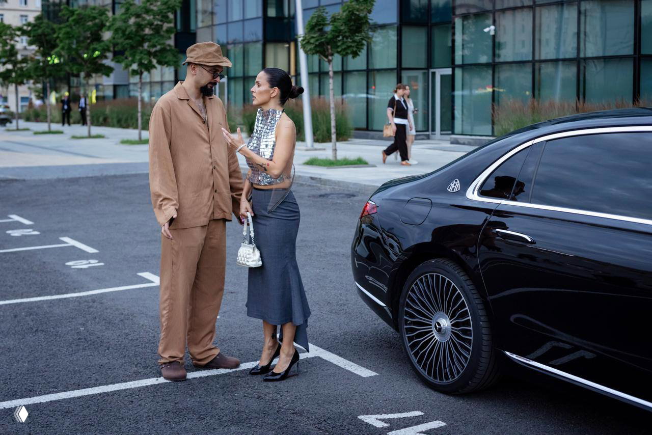 Певица ведёт беседу с мужчиной рядом с чёрным автомобилем Maybach на парковке; видны дорожная разметка, пешеходы и городская среда.