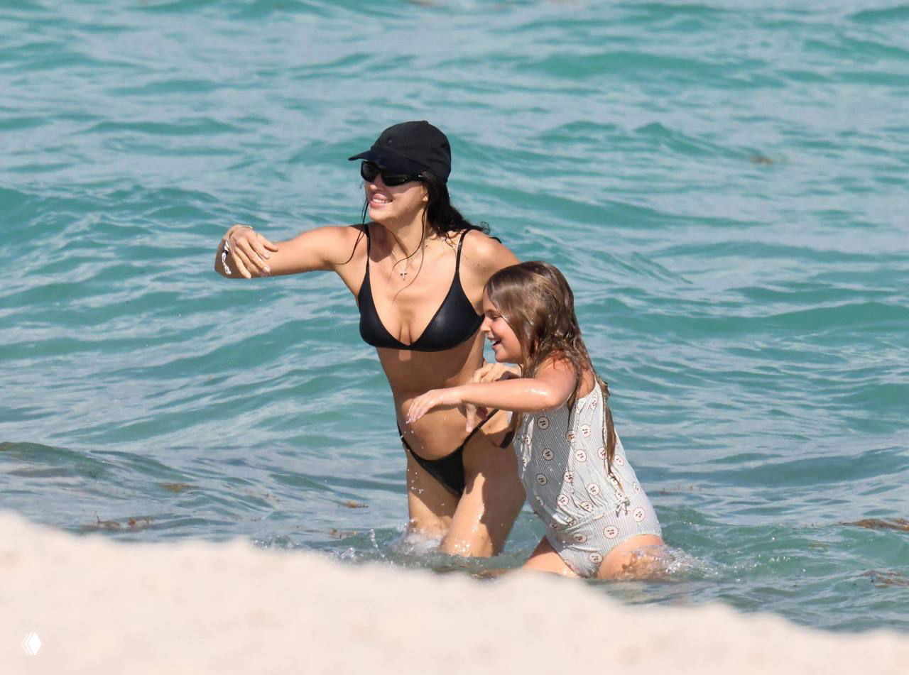 Женщина в чёрном бикини ведёт девочку за руку и вместе заходят в воду; солнечная сцена семейного пляжного отдыха у моря.