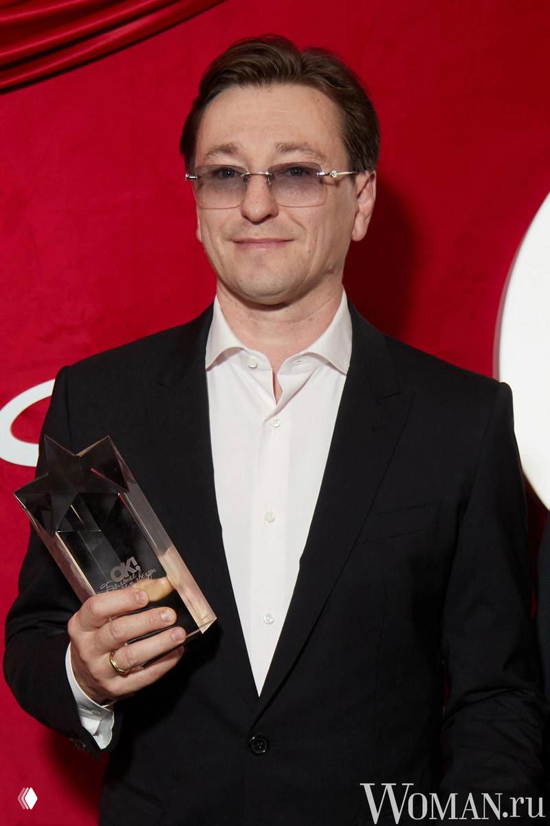 Мужчина в тёмном костюме держит статуэтку на красном фоне; портретный кадр с наградой и официальной обстановкой мероприятия.