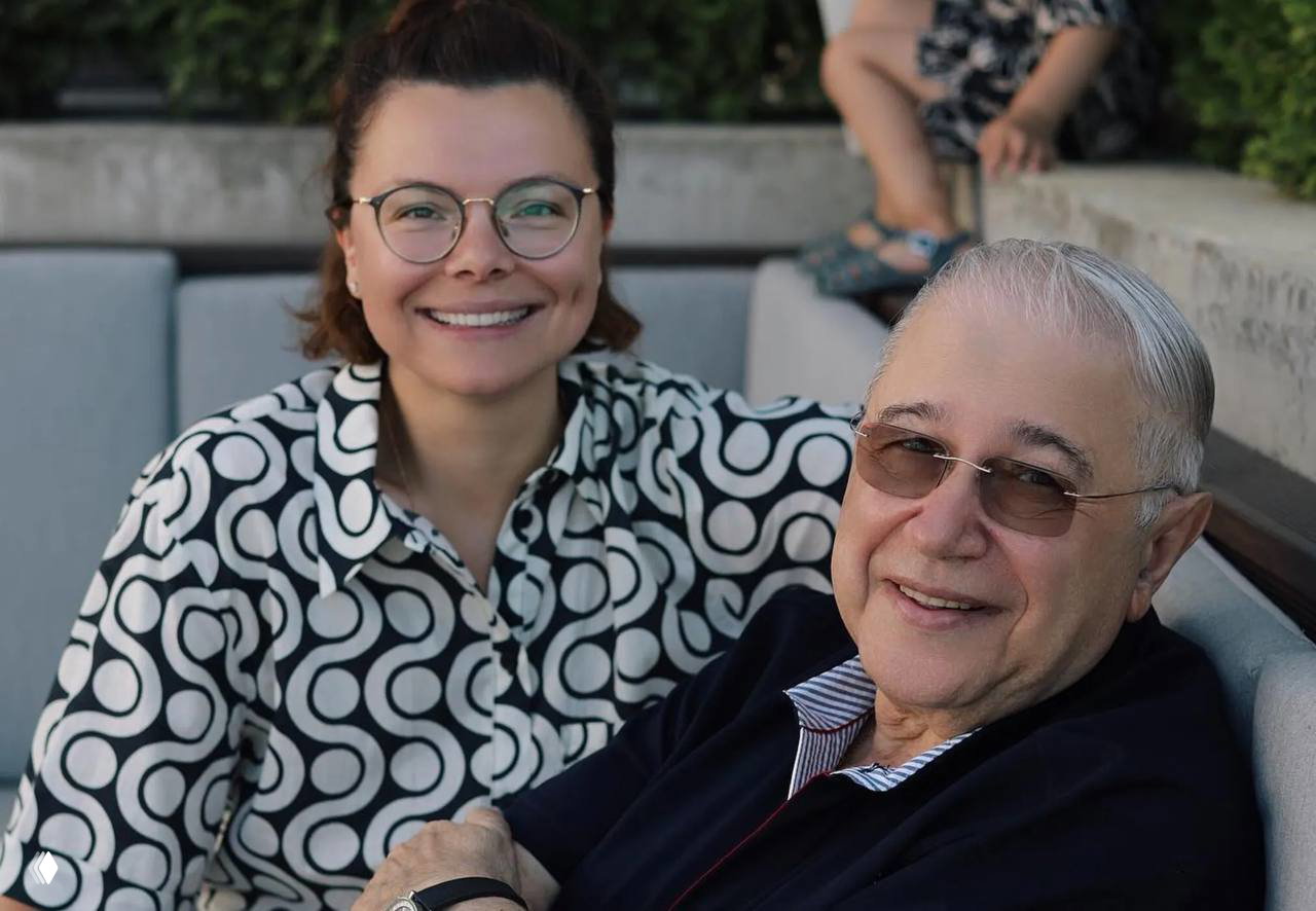 Улыбающаяся пара сидит на уличной скамье: женщина в очках и мужчина в солнцезащитных очках в непринуждённой обстановке на террасе.