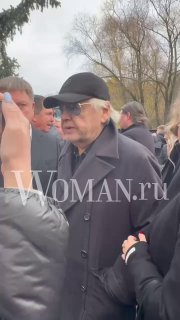 Крупный план пожилого мужчины в тёмных очках и кепке среди собравшихся — кадр с прощальной церемонии у храма.