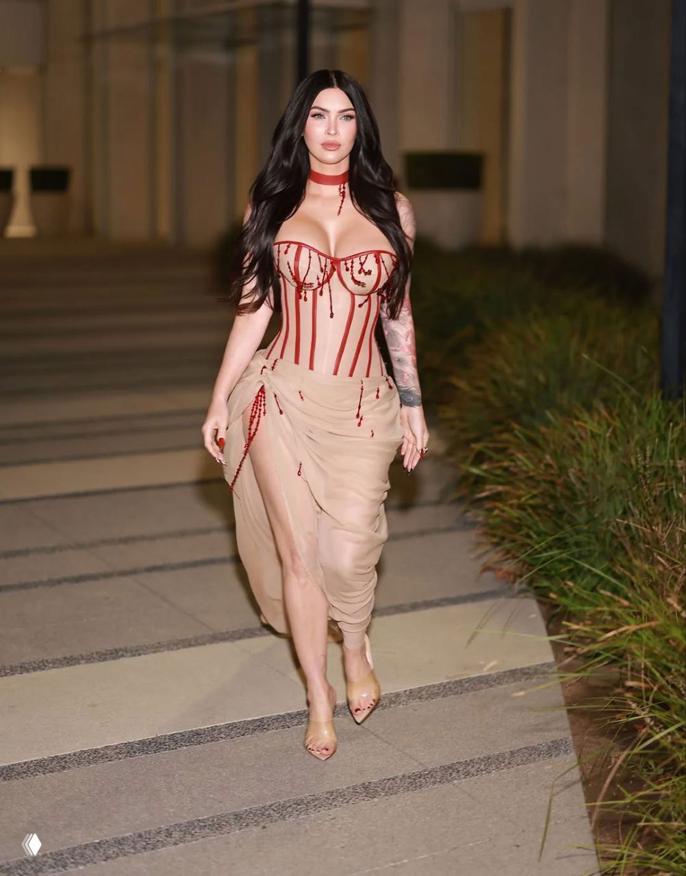 Фигура в бежевом корсете с декоративными красными бусинами на фоне архитектуры, вечерняя съёмка на улице.