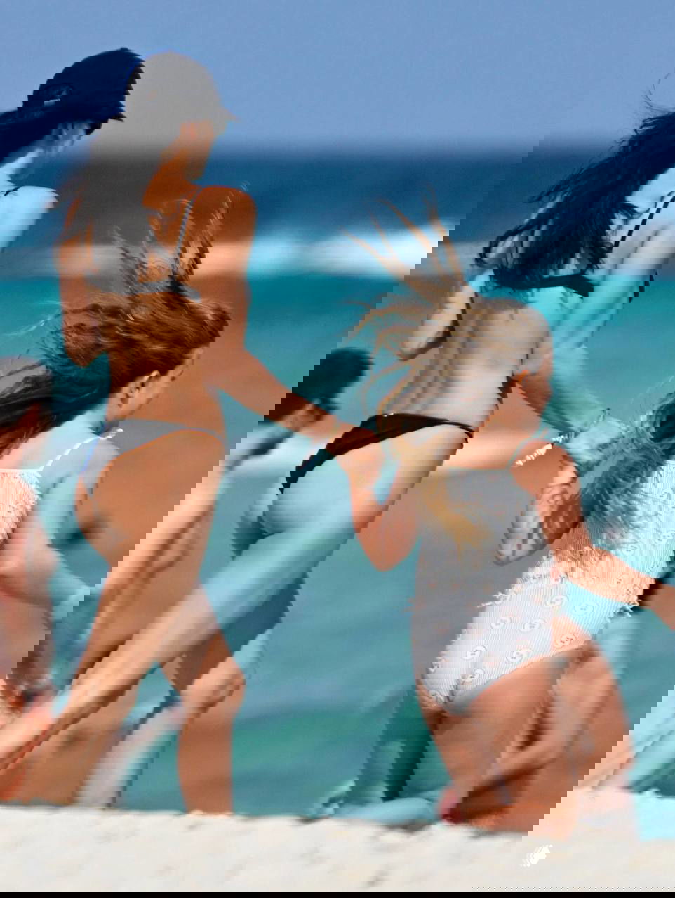 Кадр сзади: женщина в бикини и девочка в купальнике держатся за руки и идут по пляжу, ветер в волосах и волны на горизонте.