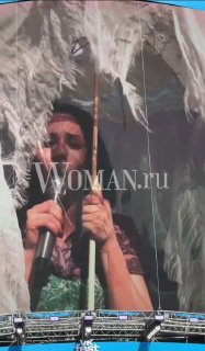 Крупный план артистки с зонтом и декоративными крыльями на фоне сцены VK FEST; видно микрофон и водяной знак Woman.ru на кадре.