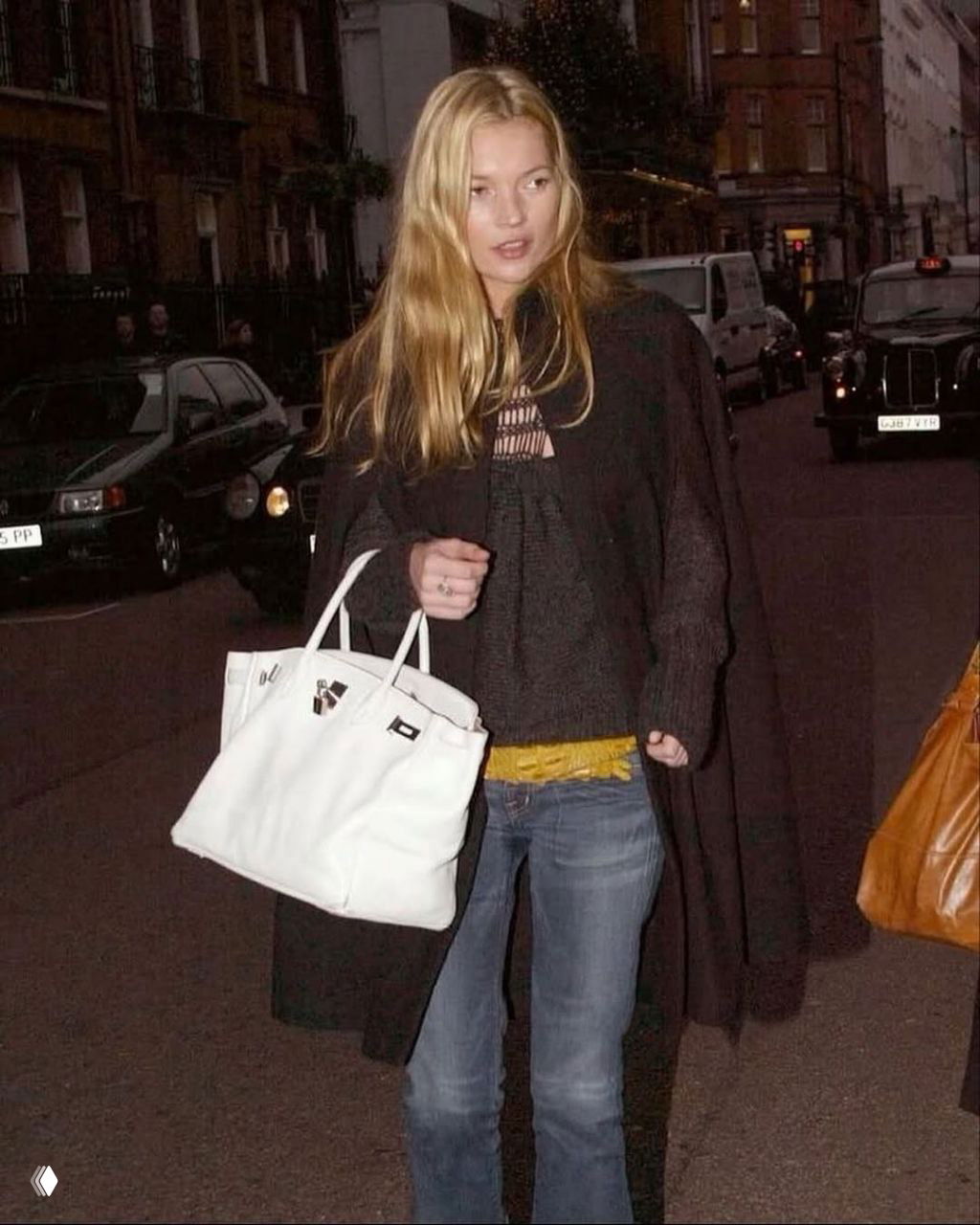 Длинное тёмное пальто с большой белой сумкой и джинсами с низкой посадкой — вечерний городской кадр с простым, но эффективным луком.