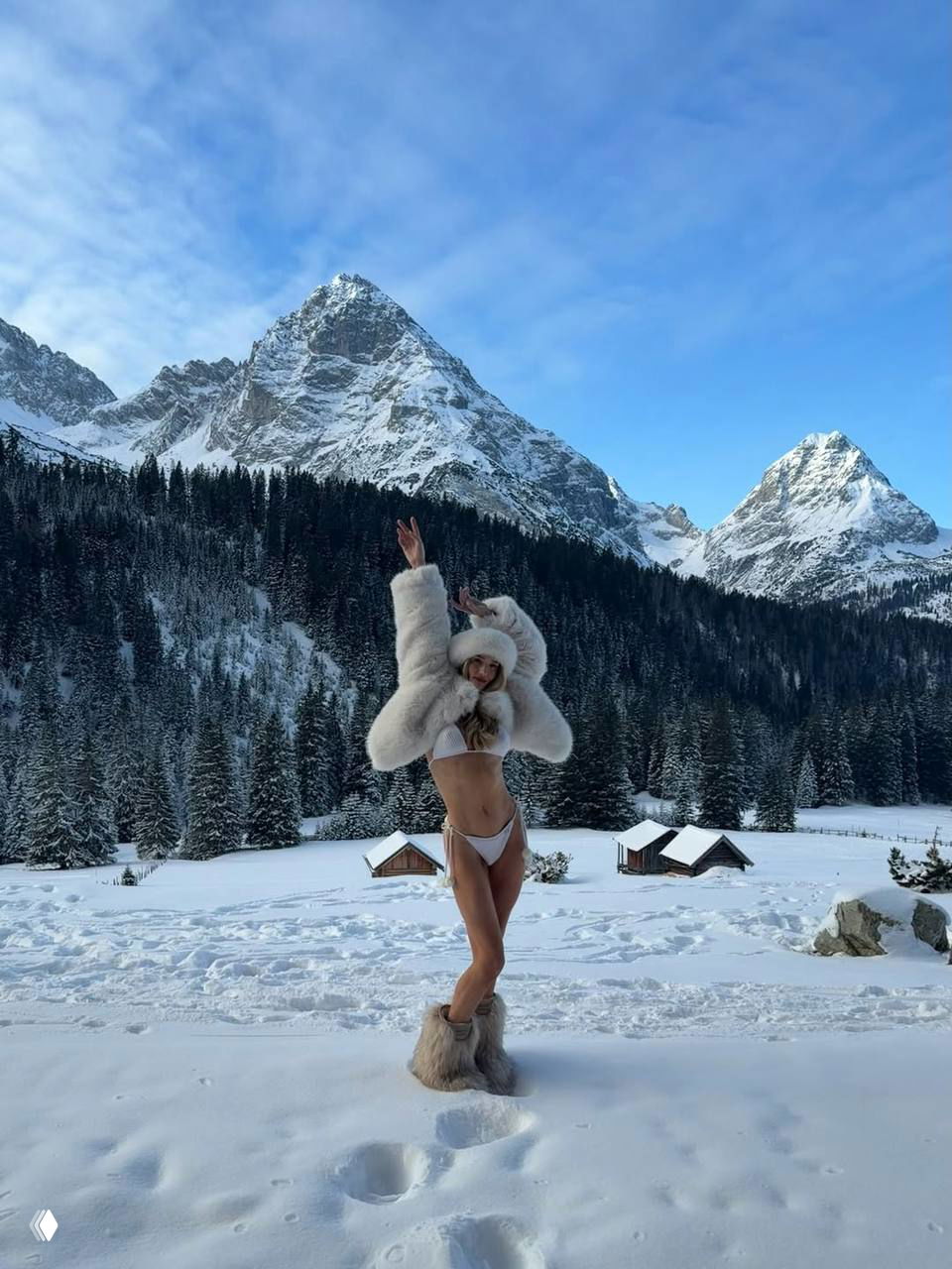 Модель в белом бикини и меховой шубе делает динамичную позу на заснеженной поляне, на заднем плане видны горы и лес.